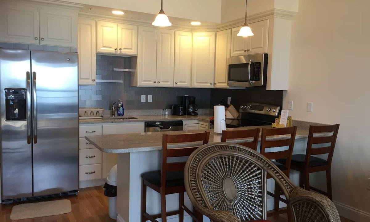 A modern kitchen with white cabinets, stainless steel appliances, and a small island with three wooden chairs. A round chair with a woven backrest is in the foreground.