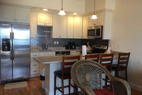 A modern kitchen with white cabinets, stainless steel appliances, and a small island with three wooden chairs. A round chair with a woven backrest is in the foreground.