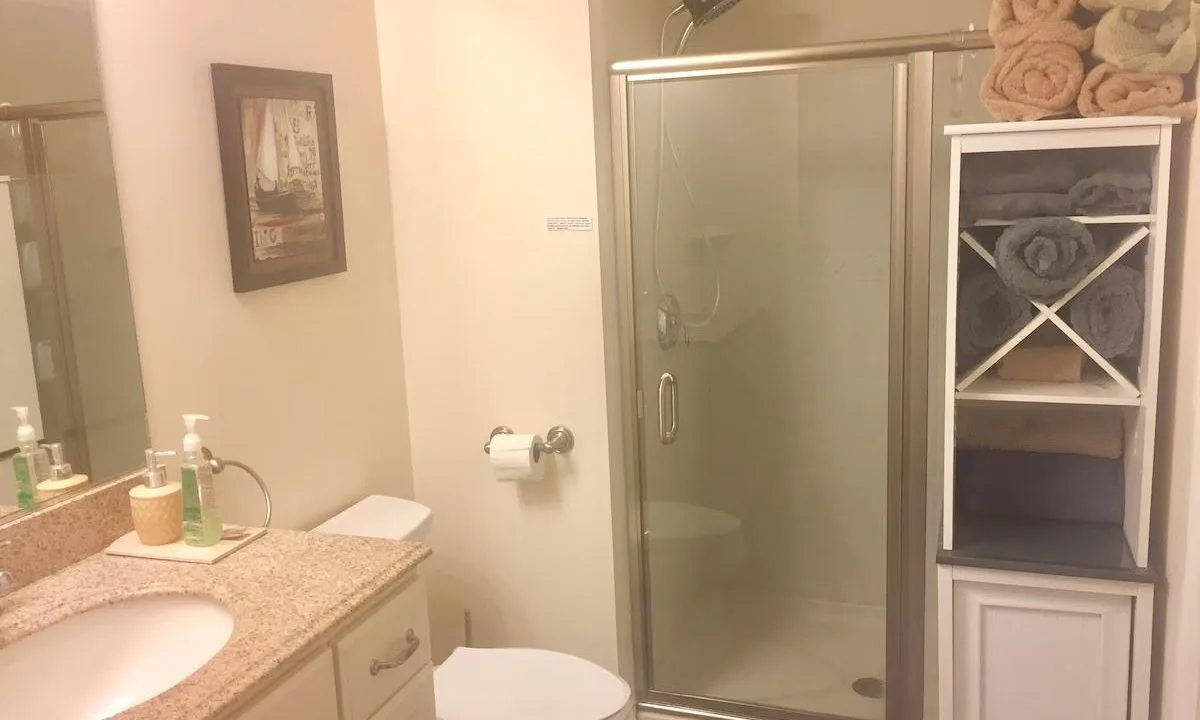 Bathroom with a shower, toilet, and sink with a granite countertop. A cabinet holds towels. A framed picture is on the wall. Toiletries include soap and a lotion bottle on the counter.