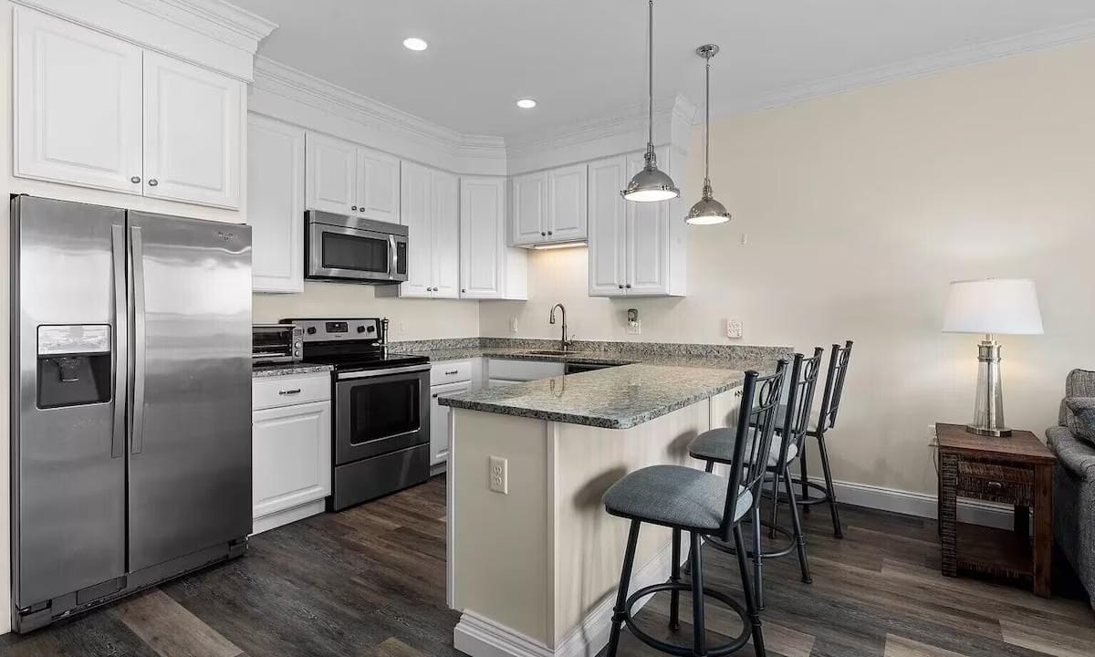 Modern kitchen with white cabinets, stainless steel appliances, granite countertops, and a breakfast bar with three metal stools on a dark wood floor.