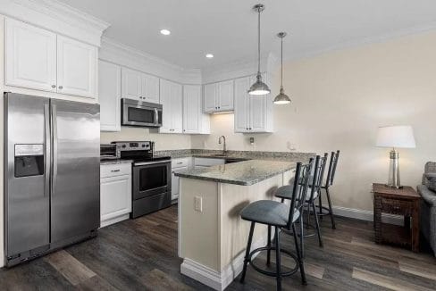 Modern kitchen with white cabinets, stainless steel appliances, granite countertops, and a breakfast bar with three metal stools on a dark wood floor.