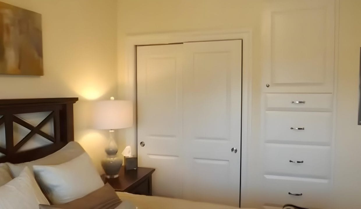 Bedroom with a bed, nightstand, table lamp, and closed sliding closet doors. A set of built-in drawers is on the wall, and artwork hangs above the bed.