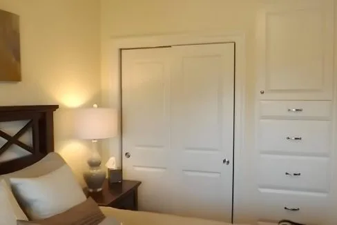 Bedroom with a bed, nightstand, table lamp, and closed sliding closet doors. A set of built-in drawers is on the wall, and artwork hangs above the bed.