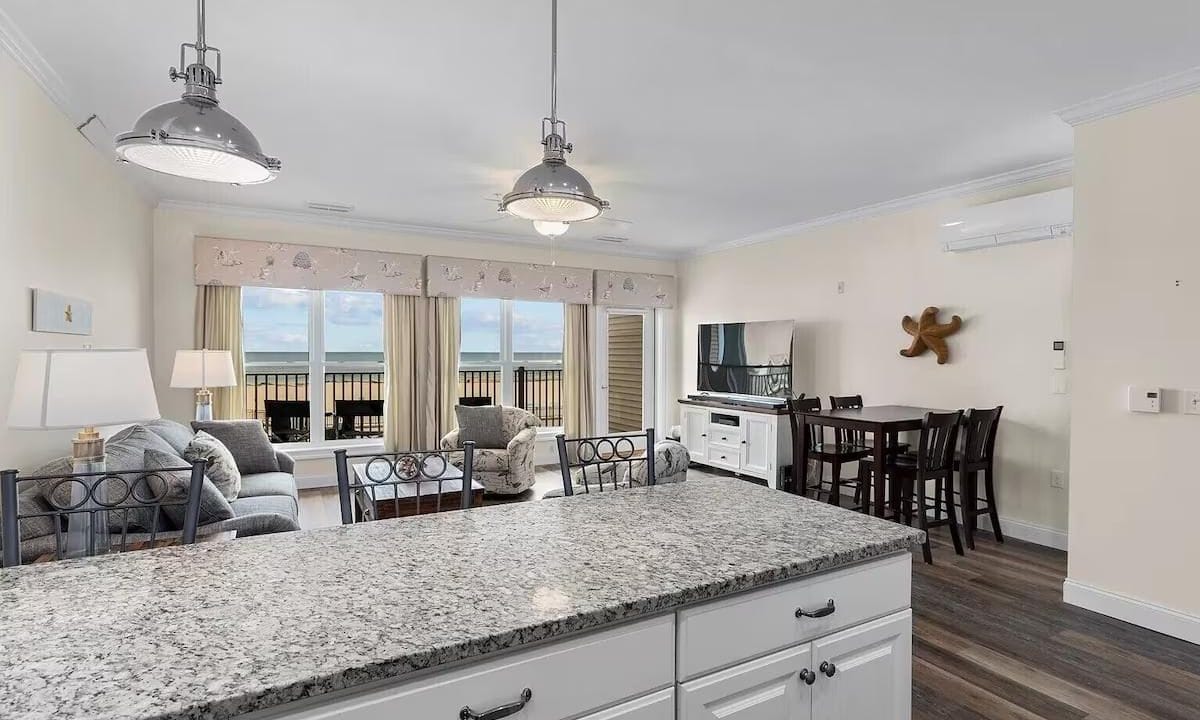 Bright open-plan living room and kitchen with granite countertops, sofa, TV, and dining table. Large windows offer a view of the balcony and ocean. Industrial-style pendant lights overhead.