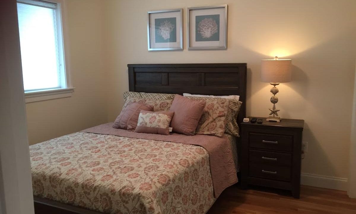 A bedroom with a wooden bed, floral-patterned bedding, two framed artworks above the headboard, a nightstand with a lamp, and a window on the left.