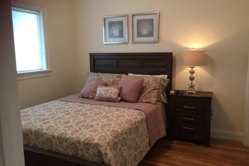 A bedroom with a wooden bed, floral-patterned bedding, two framed artworks above the headboard, a nightstand with a lamp, and a window on the left.