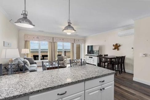 Bright open-plan living room and kitchen with granite countertops, sofa, TV, and dining table. Large windows offer a view of the balcony and ocean. Industrial-style pendant lights overhead.