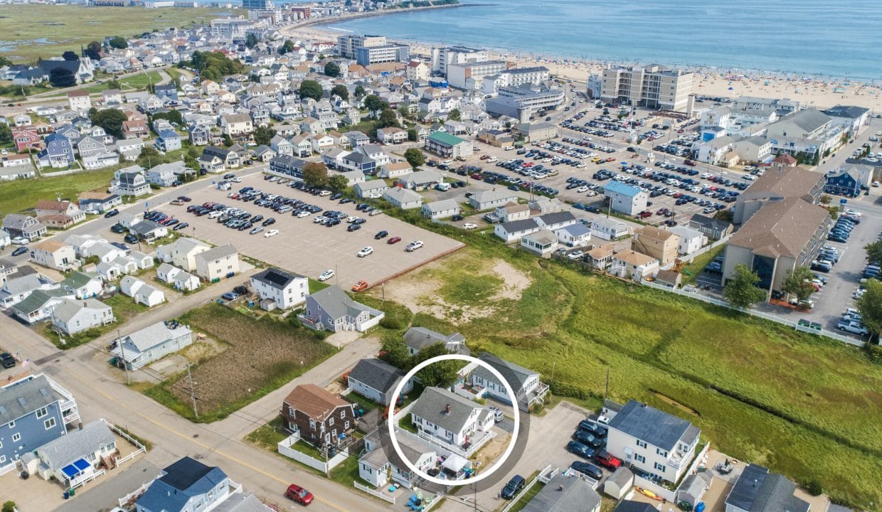 Aerial view of a coastal town with residential houses, parking lots, and beach in the background. A white circle highlights a specific house.