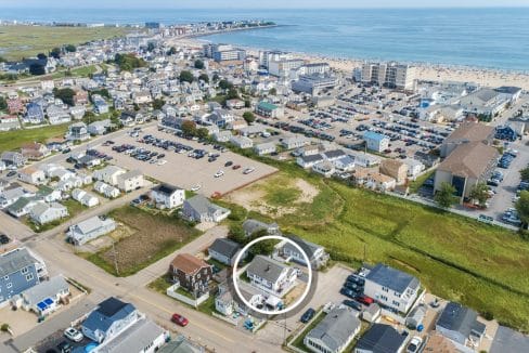 Aerial view of a coastal town with residential houses, parking lots, and beach in the background. A white circle highlights a specific house.