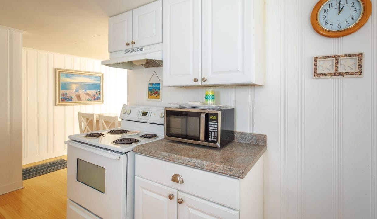 A compact kitchen features white cabinets, an electric stove, a microwave, and a countertop. A wall clock and framed pictures hang above the counter. Dining table visible in the next room.