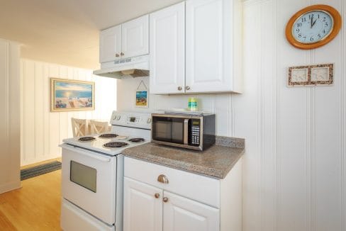 A compact kitchen features white cabinets, an electric stove, a microwave, and a countertop. A wall clock and framed pictures hang above the counter. Dining table visible in the next room.