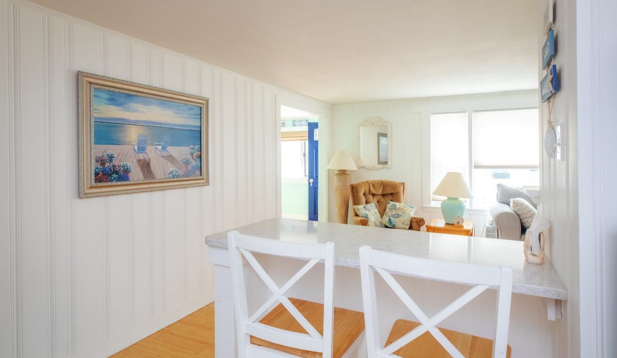 Bright coastal-themed living area with white walls, decorative ocean painting, two white bar stools, and cozy seating near large windows.