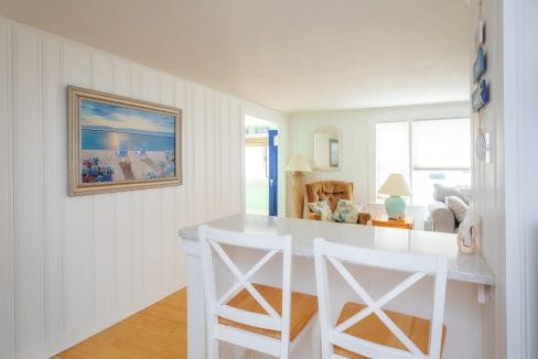 Bright coastal-themed living area with white walls, decorative ocean painting, two white bar stools, and cozy seating near large windows.