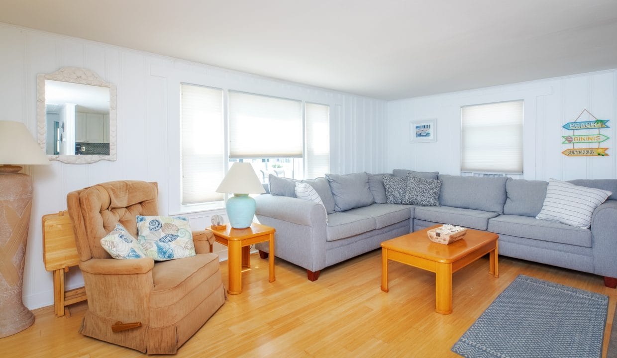 Cozy living room with a gray sectional sofa, a beige armchair, wooden coffee table, and soft lighting. Blue rug on light wood flooring under a large window.