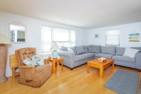 Cozy living room with a gray sectional sofa, a beige armchair, wooden coffee table, and soft lighting. Blue rug on light wood flooring under a large window.