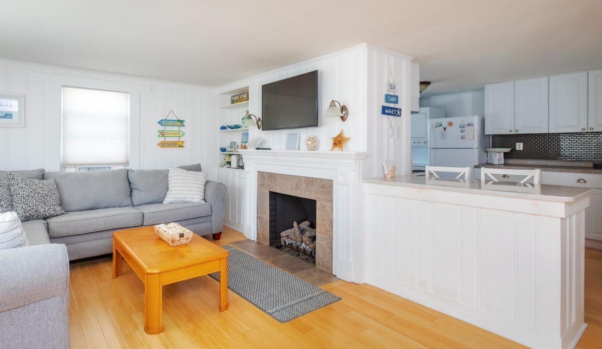 Cozy living room with a gray sofa, wooden coffee table, and a fireplace. White walls and cabinets in the background with a TV mounted above the mantel. .