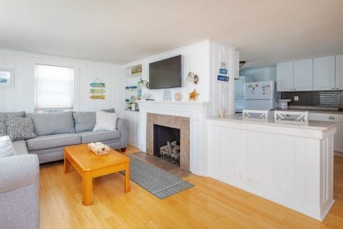 Cozy living room with a gray sofa, wooden coffee table, and a fireplace. White walls and cabinets in the background with a TV mounted above the mantel. .