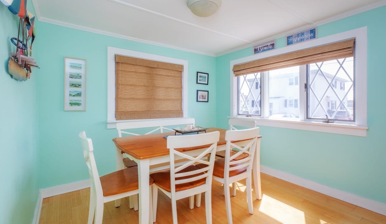 Dining room with light green walls, wooden table, and white chairs. Two large windows with blinds let in natural light. Framed pictures adorn the walls.