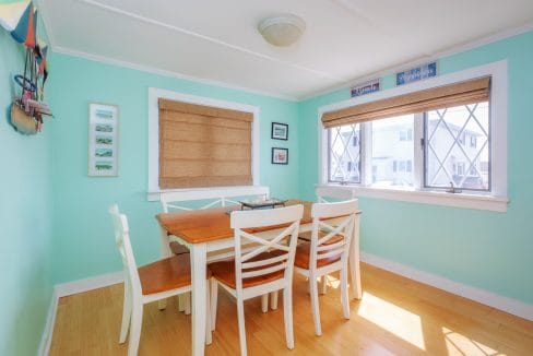 Dining room with light green walls, wooden table, and white chairs. Two large windows with blinds let in natural light. Framed pictures adorn the walls.