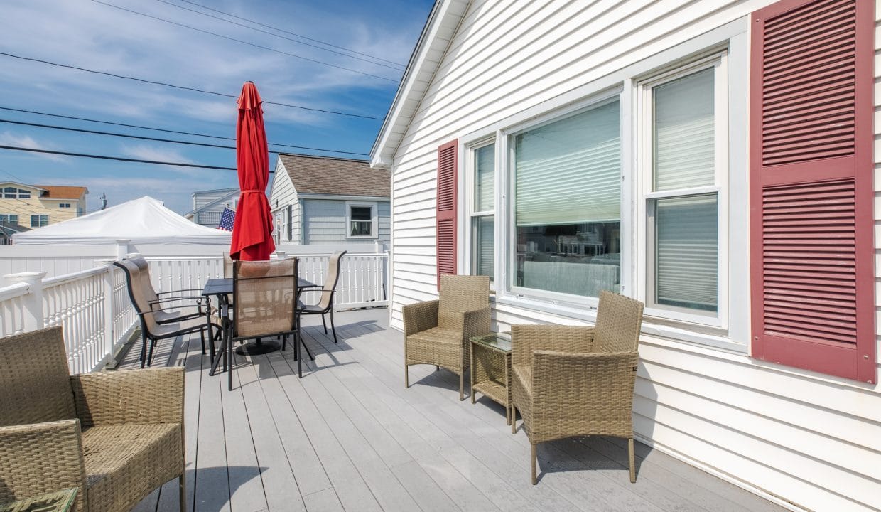 A rooftop patio with wicker furniture, a table, a red umbrella, and a white house exterior.