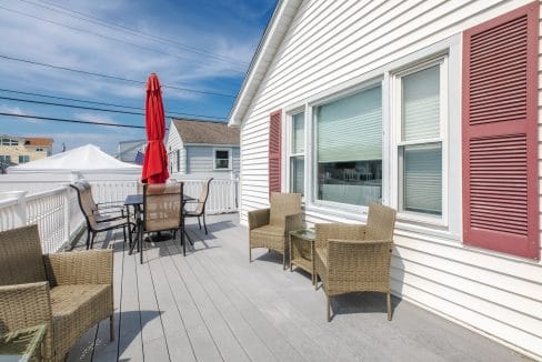 A rooftop patio with wicker furniture, a table, a red umbrella, and a white house exterior.