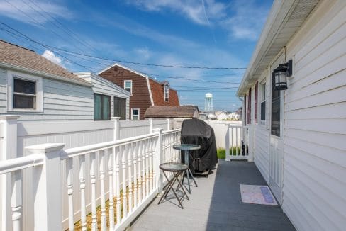 A white fenced deck with a grill and two chairs under a blue sky. Houses and a water tower are visible in the background.