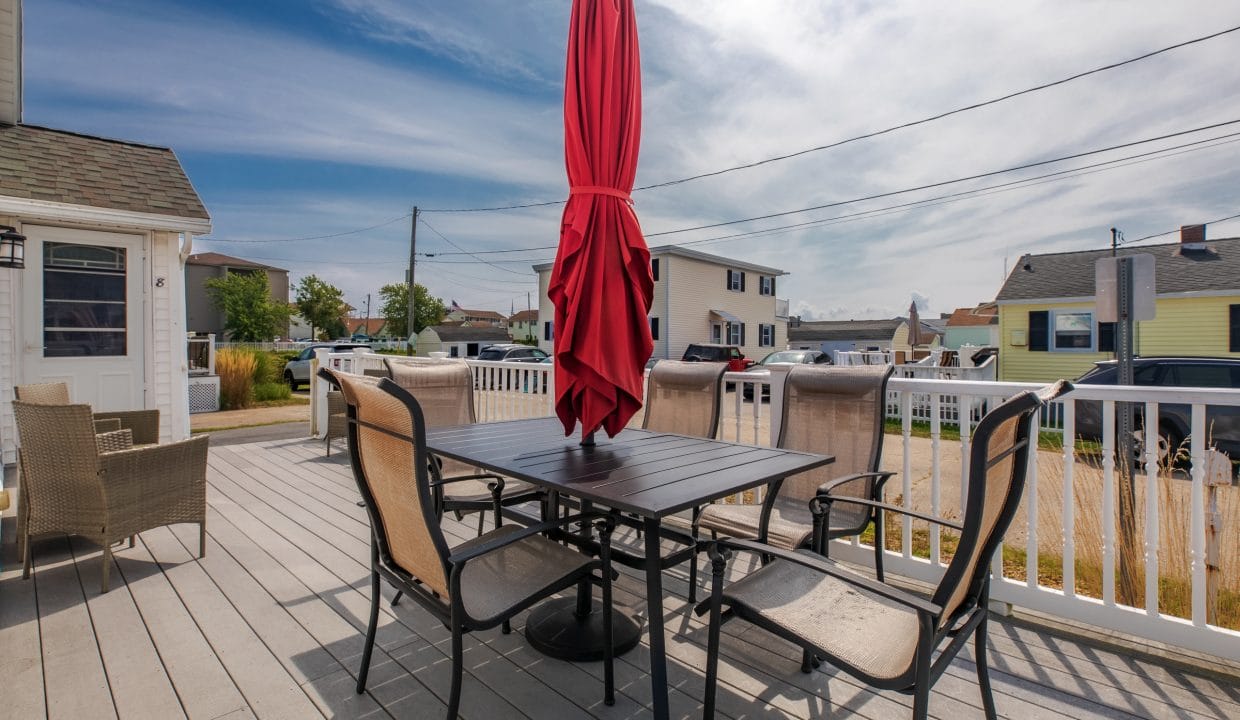 A patio with a table and six chairs under a closed red umbrella, surrounded by a white railing, with houses and a clear sky in the background.