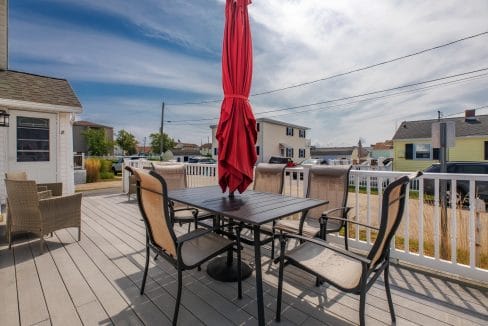 A patio with a table and six chairs under a closed red umbrella, surrounded by a white railing, with houses and a clear sky in the background.