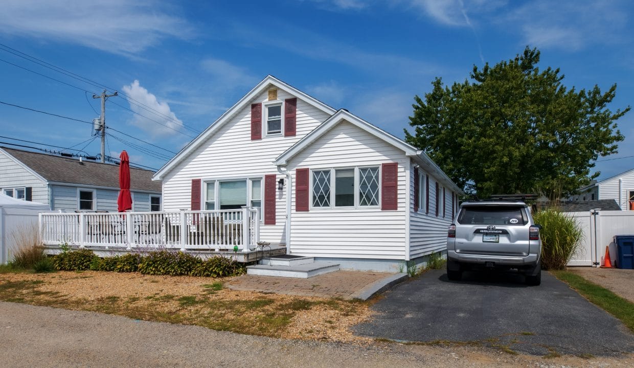 A small white house with red shutters, a porch with a red umbrella, and an SUV parked in the driveway under a blue sky.