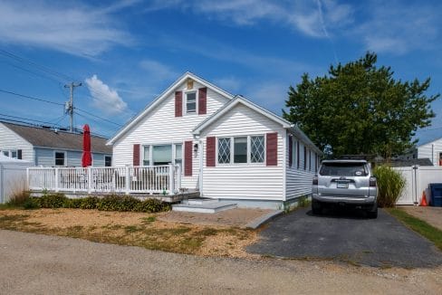 A small white house with red shutters, a porch with a red umbrella, and an SUV parked in the driveway under a blue sky.