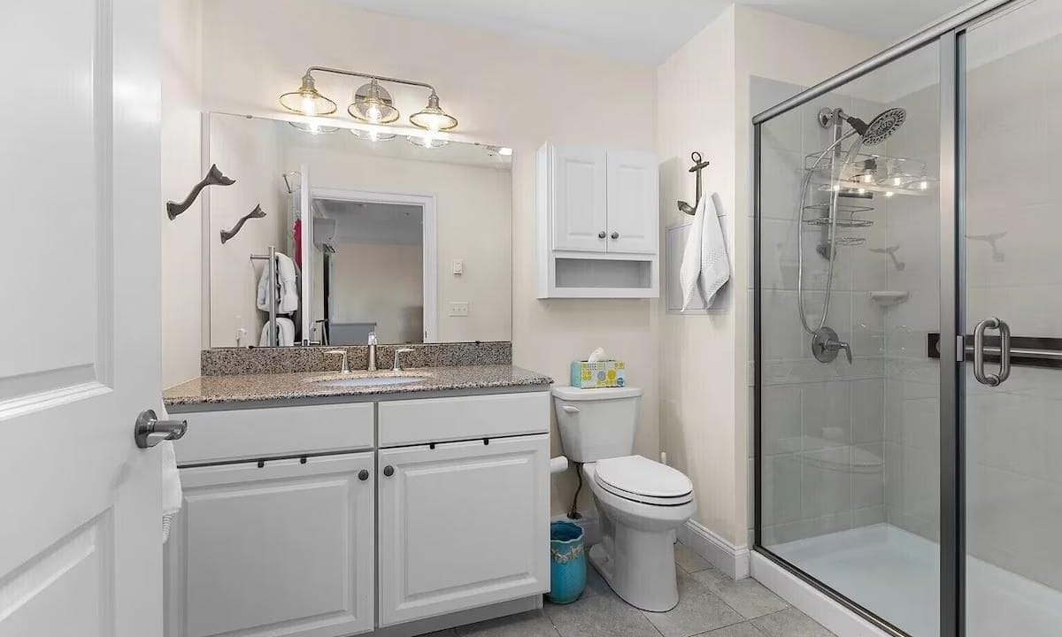 Modern bathroom with a granite countertop, large mirror, dual sinks, toilet, and glass-enclosed shower. Cabinets and towel hangers are mounted on the cream-colored walls.