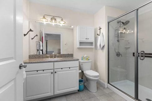 Modern bathroom with a granite countertop, large mirror, dual sinks, toilet, and glass-enclosed shower. Cabinets and towel hangers are mounted on the cream-colored walls.