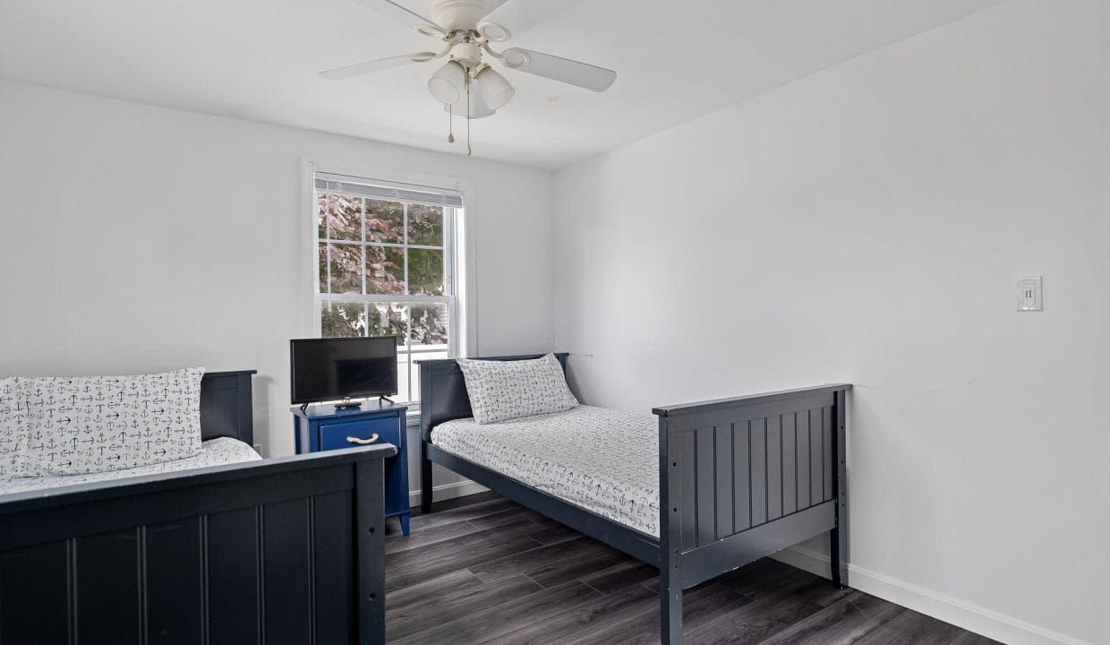 A small bedroom with two single black beds, a blue nightstand, a small TV, and a ceiling fan. The room has a window with a view of trees and features dark wooden flooring.