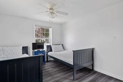 A small bedroom with two single black beds, a blue nightstand, a small TV, and a ceiling fan. The room has a window with a view of trees and features dark wooden flooring.