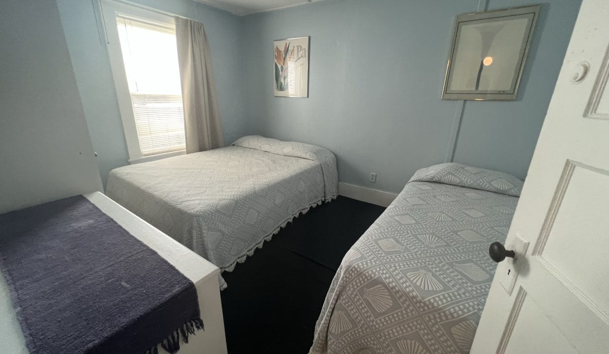 A room with two beds, each covered in patterned bedspreads, a window with a curtain, and a framed picture on the wall.