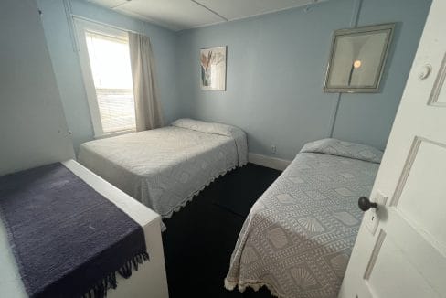 A room with two beds, each covered in patterned bedspreads, a window with a curtain, and a framed picture on the wall.