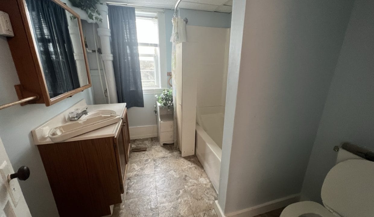 Bathroom with a sink, mirror, shower-tub combo, toilet, and window with curtains. There are plants near the window and a cabinet below the sink. The flooring is tiled.