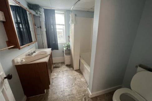 Bathroom with a sink, mirror, shower-tub combo, toilet, and window with curtains. There are plants near the window and a cabinet below the sink. The flooring is tiled.