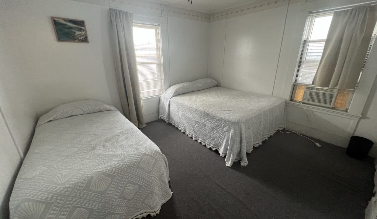 A simple bedroom with two beds, each covered with white patterned bedspreads. There are two windows with curtains and a ceiling fan above. A small framed picture hangs on the wall.