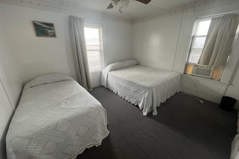 A simple bedroom with two beds, each covered with white patterned bedspreads. There are two windows with curtains and a ceiling fan above. A small framed picture hangs on the wall.