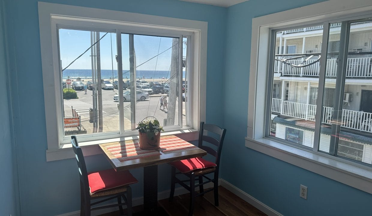 Small dining table with two red chairs by large windows, overlooking a street and parking area by the ocean. Blue walls and wooden floor. A potted plant sits on the table.