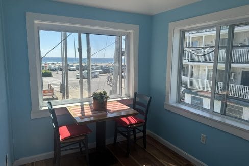 Small dining table with two red chairs by large windows, overlooking a street and parking area by the ocean. Blue walls and wooden floor. A potted plant sits on the table.