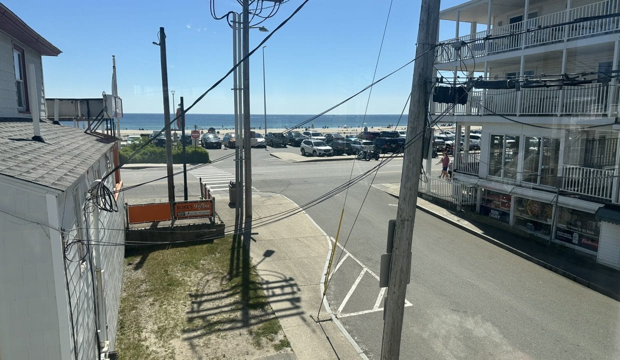 View from a window showing a street intersection near a beach, with parked cars, utility poles, and a multi-story building with balconies under a clear blue sky.