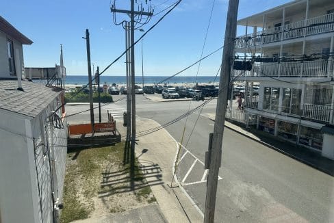 View from a window showing a street intersection near a beach, with parked cars, utility poles, and a multi-story building with balconies under a clear blue sky.