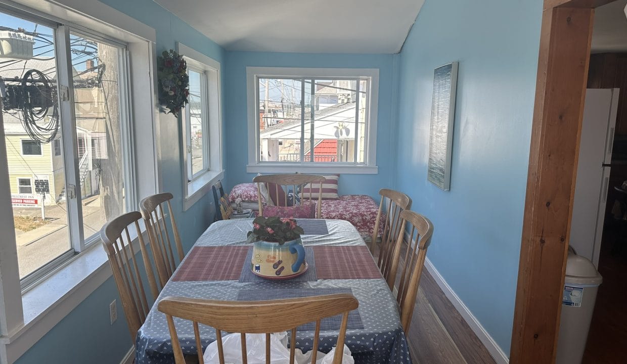 A narrow dining area with a table set for six, featuring a floral centerpiece. Bright blue walls, large windows, and a small bed in the corner. Urban view outside.