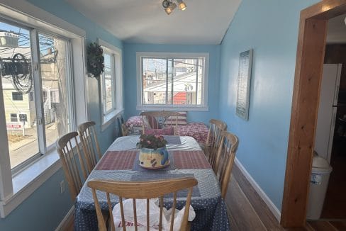 A narrow dining area with a table set for six, featuring a floral centerpiece. Bright blue walls, large windows, and a small bed in the corner. Urban view outside.