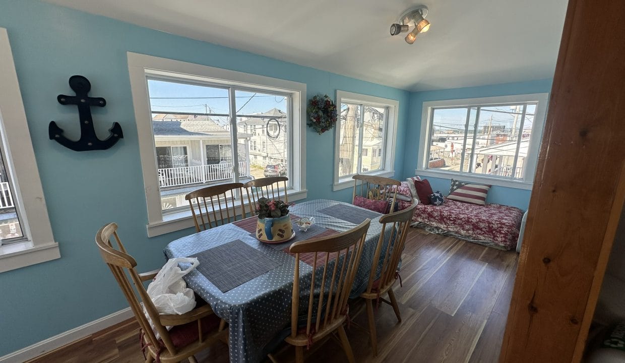 Dining area with a wooden table and chairs, blue walls, nautical decor, and large windows. Red-patterned cushions on a bench. Sunlight streaming in.