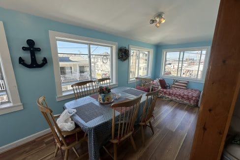 Dining area with a wooden table and chairs, blue walls, nautical decor, and large windows. Red-patterned cushions on a bench. Sunlight streaming in.