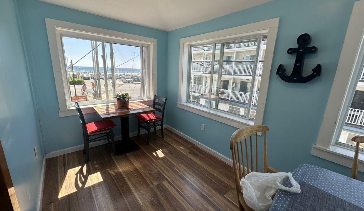 A bright dining area with light blue walls, two windows, a small table with red chairs, a large anchor wall decoration, and wood flooring. A view of parked cars and a building is outside.