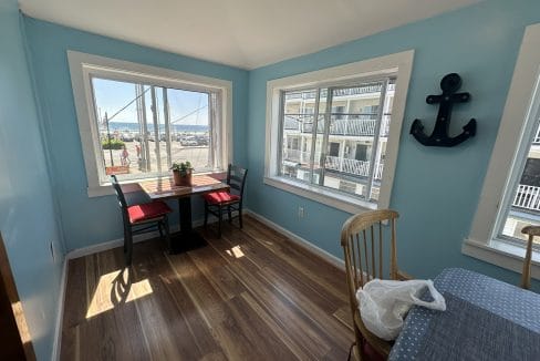 A bright dining area with light blue walls, two windows, a small table with red chairs, a large anchor wall decoration, and wood flooring. A view of parked cars and a building is outside.